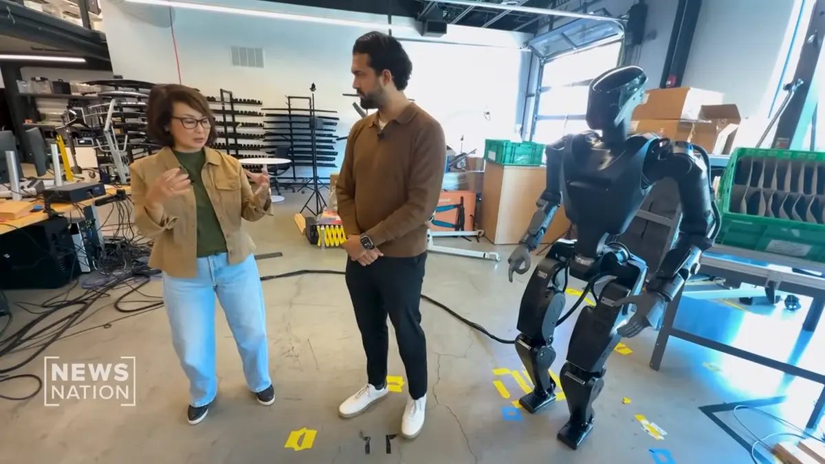 Foundation Robotics CEO Sankaet Pathak stands beside a Phantom humanoid robot while speaking with NewsNation reporter Nancy Loo during a television segment about the company's military ambitions and production goals.