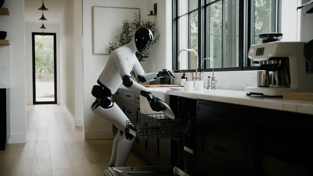 A Figure 03 humanoid robot stands in a kitchen, holding a plate as it carefully places it into the lower rack of an open dishwasher.