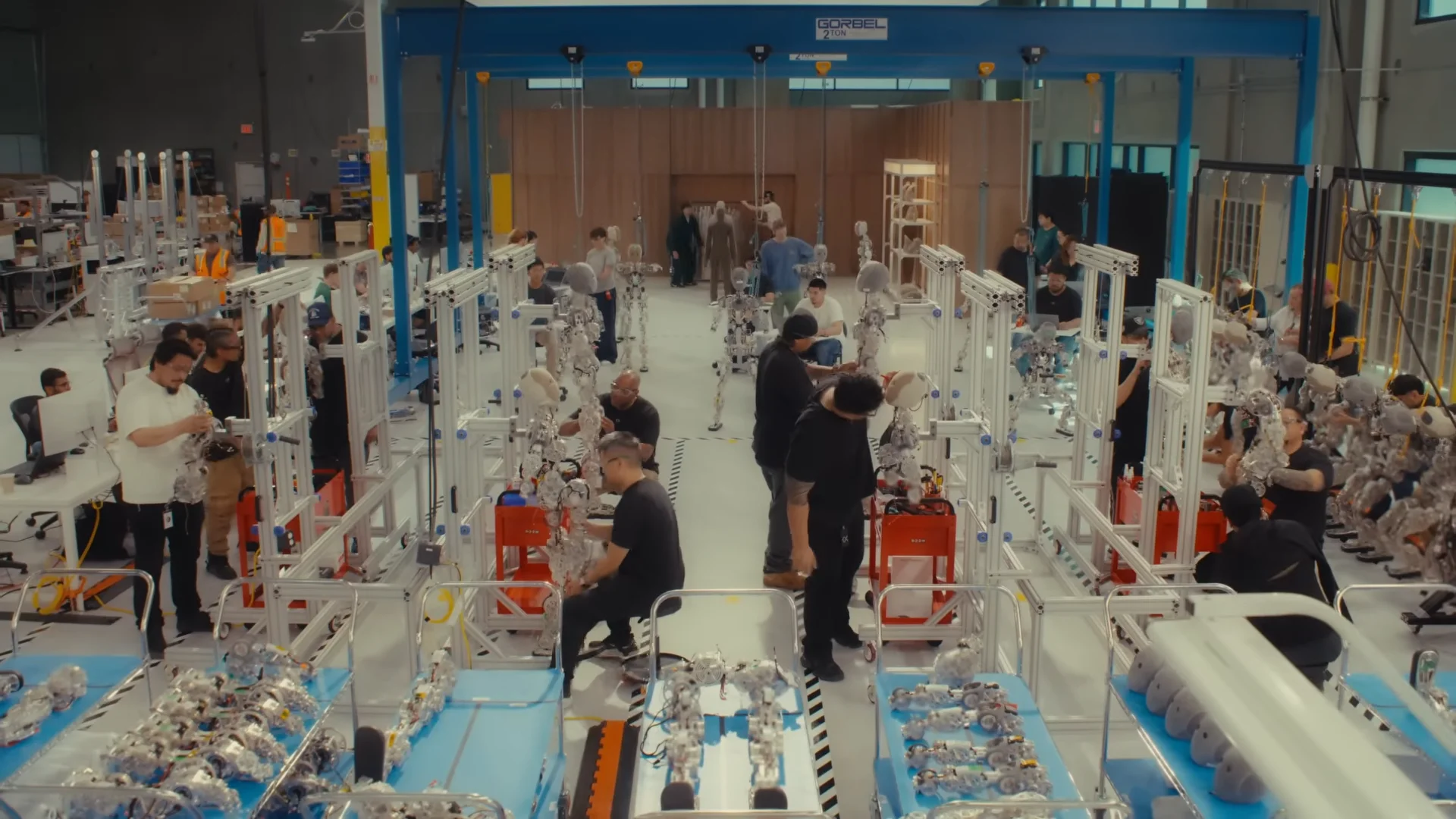 A high-angle wide shot of a brightly lit factory floor where numerous technicians in black shirts work at assembly stations equipped with silver metallic frames and blue component carts under a large blue overhead crane structure.