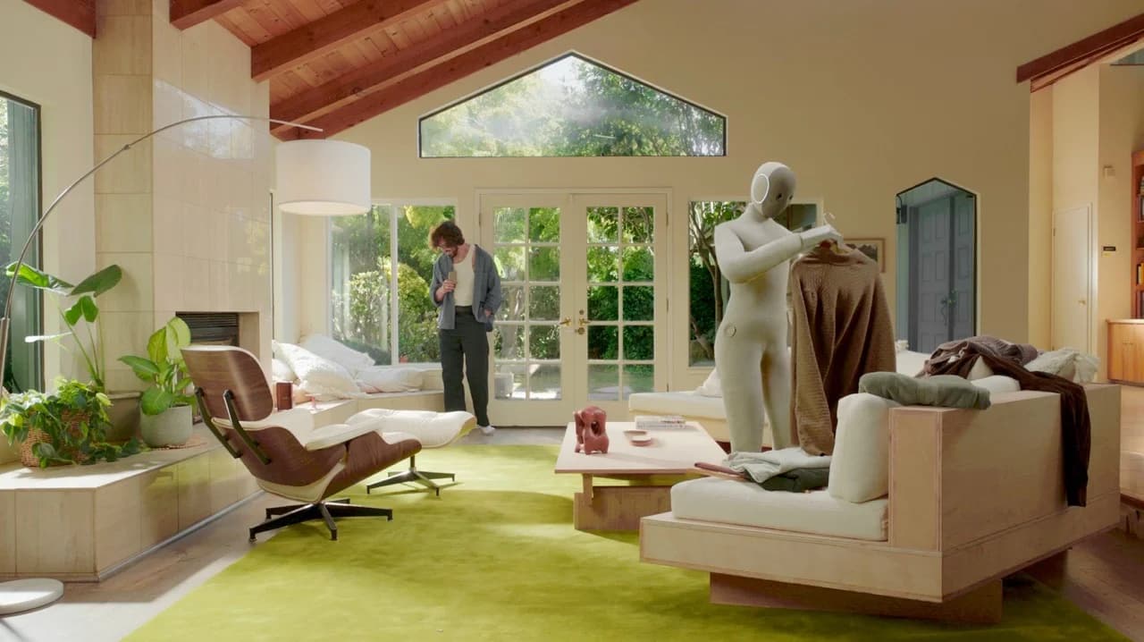 A soft-bodied, white NEO humanoid robot stands in a sunlit, modern living room, holding a brown sweater on a wooden hanger. A human man stands in the background near glass doors, casually observing.
