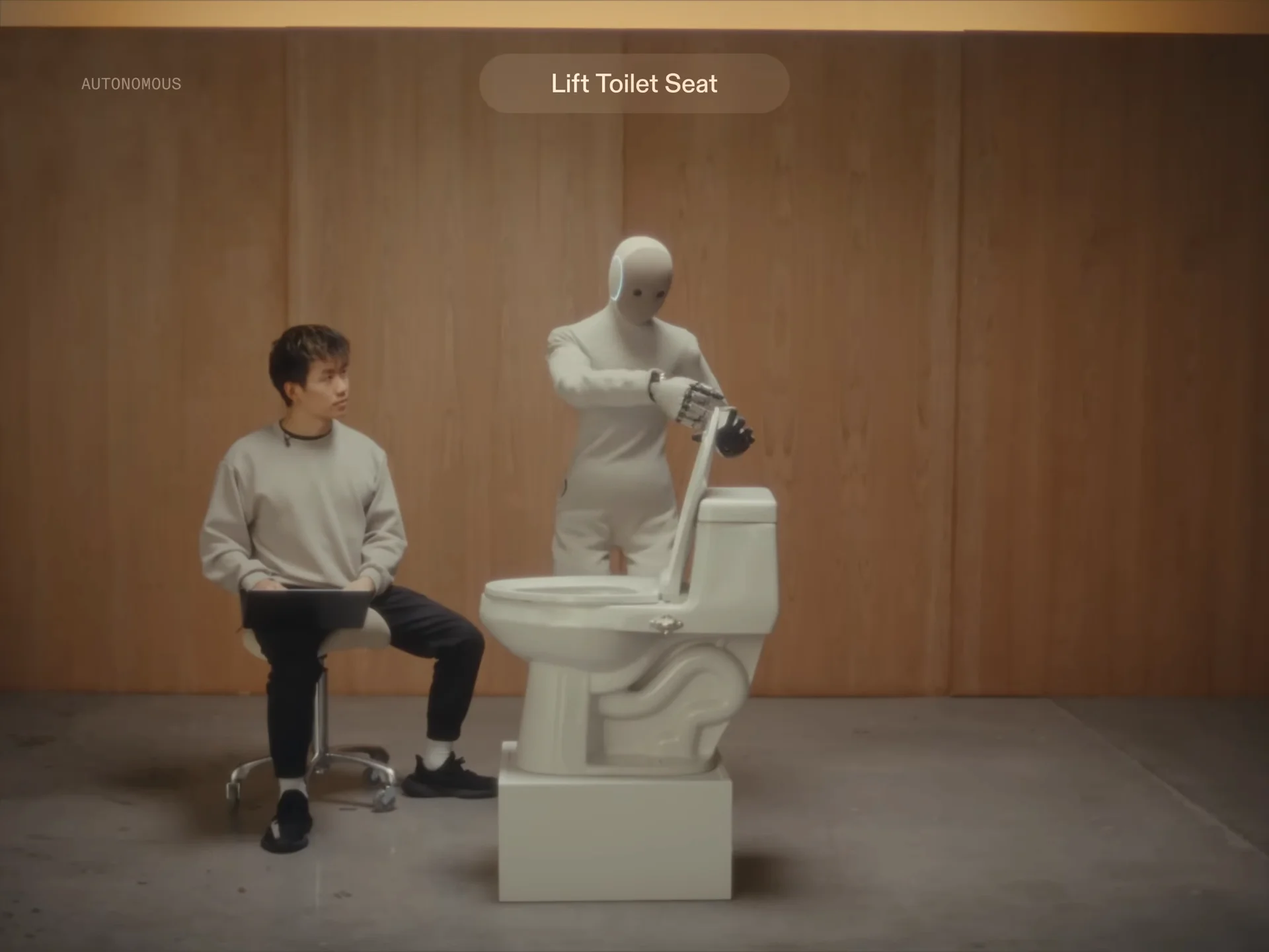 A white NEO humanoid robot standing in front of a white toilet, reaching out to lift the toilet seat while a human sits nearby watching.