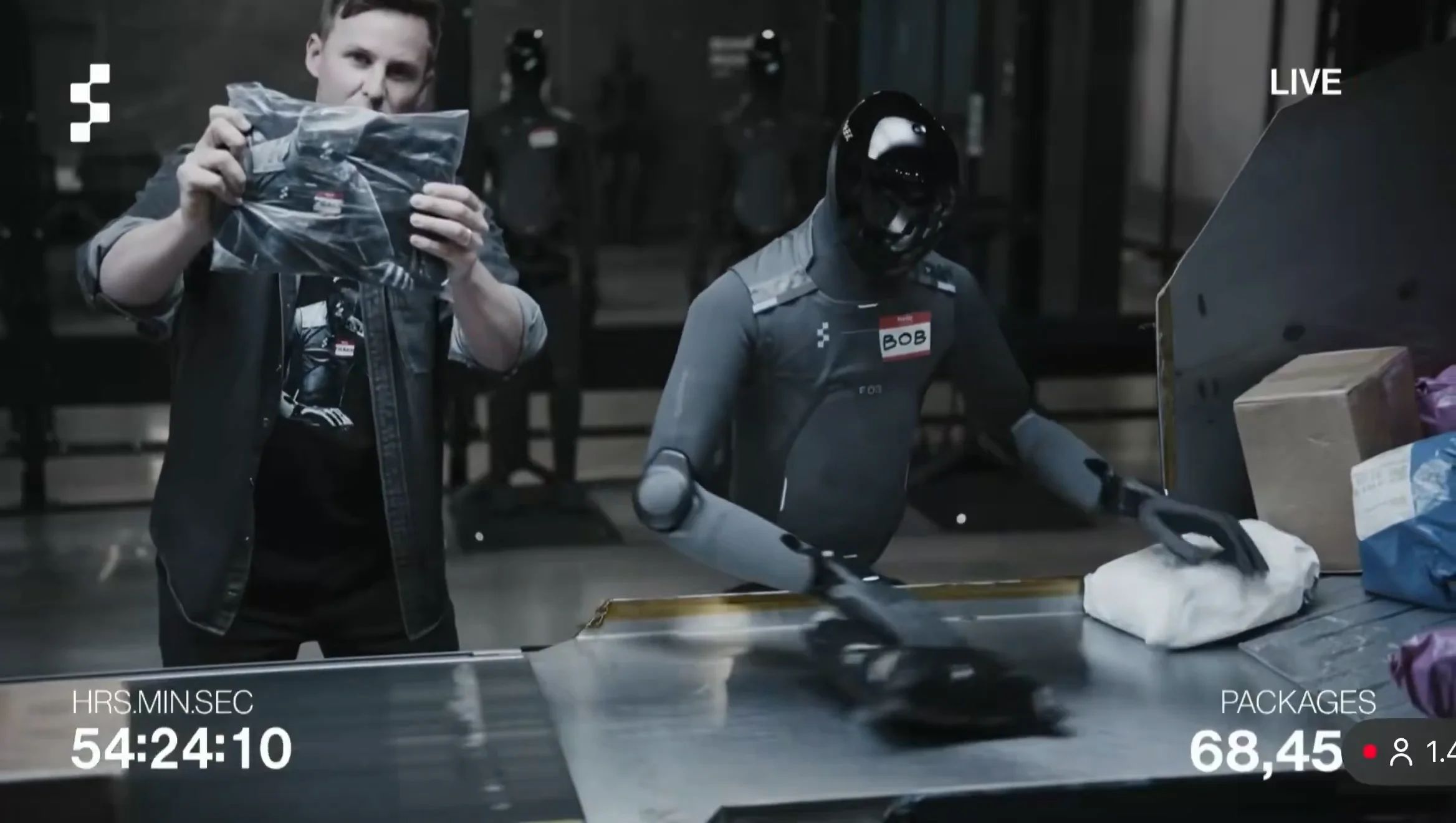 A screenshot from a Figure AI live video feed showing CEO Brett Adcock on the left holding up a black t-shirt enclosed in a clear plastic bag with a barcode tag. To his right, a charcoal-grey Figure 03 humanoid robot with a red 'BOB' name tag reaches for a package on a metal conveyor belt. Video overlays at the bottom display a runtime of 54:24:10 and a package count of 68,45X.