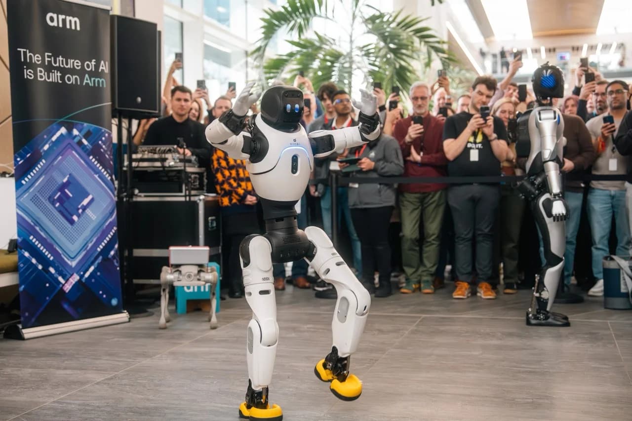 A compact white and black Lingxi X2 humanoid robot with yellow feet strikes a dynamic pose in the center of a crowded room at Arm Holdings’ UK headquarters. Behind it, a taller silver and black Expedition A2 humanoid robot stands near a large 'arm' banner, while a small D1 quadruped robot rests on a stand in the background. 