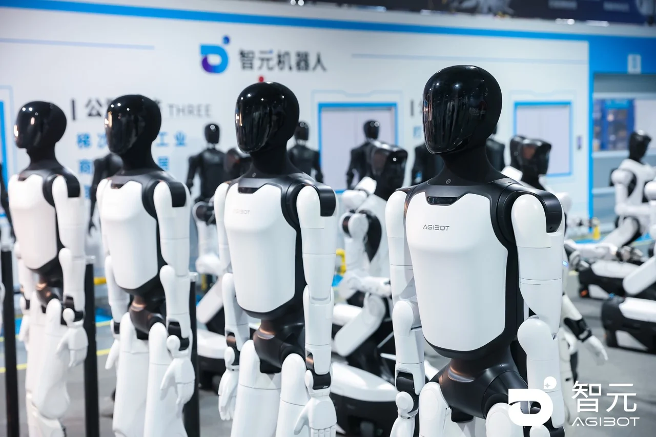 A row of several AgiBot A3 humanoid robots stands in profile in a clean, brightly lit industrial facility. The robots feature sleek black heads and white torso panels with black joints. In the background, the AgiBot logo and Chinese branding are visible on a white and blue wall.