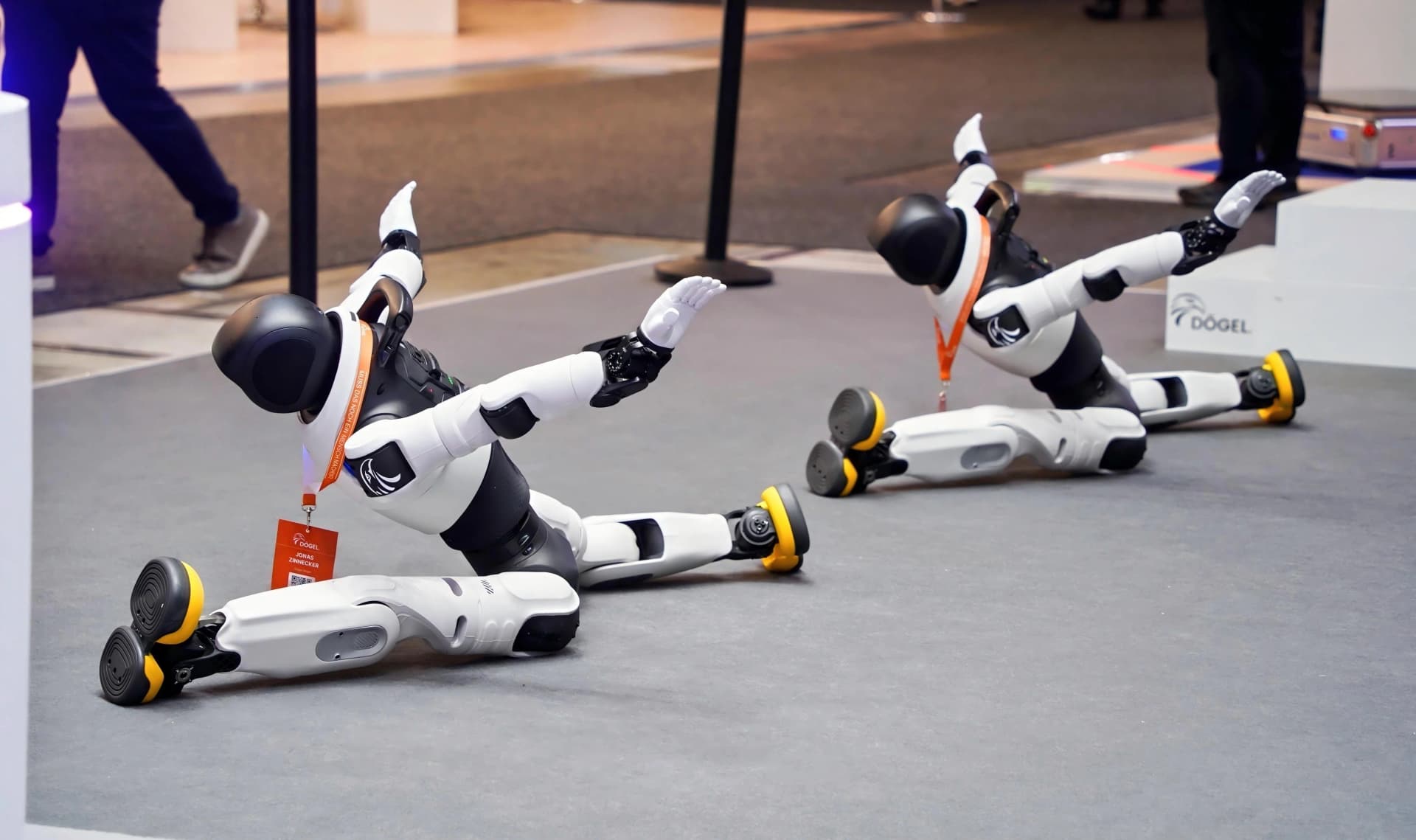 Two black and white AGIBOT X2 humanoid robots with yellow soles performing synchronized side splits and expressive dance poses on a grey stage floor at IFA 2026.