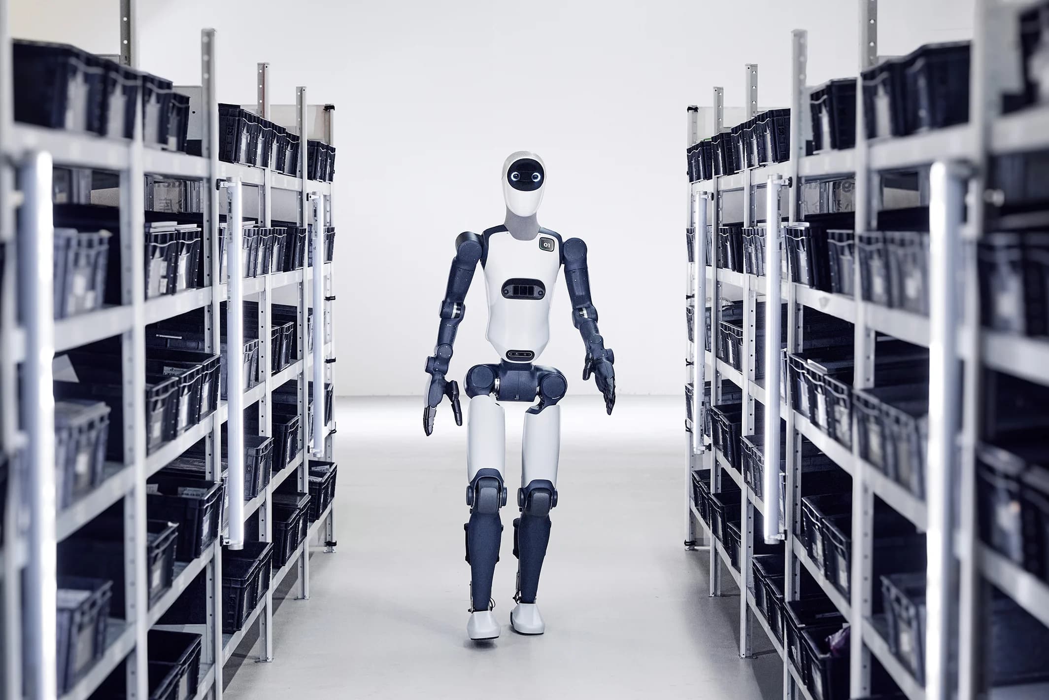 The bipedal Agile ONE humanoid robot walking down a narrow aisle between shelving units in a logistics facility.