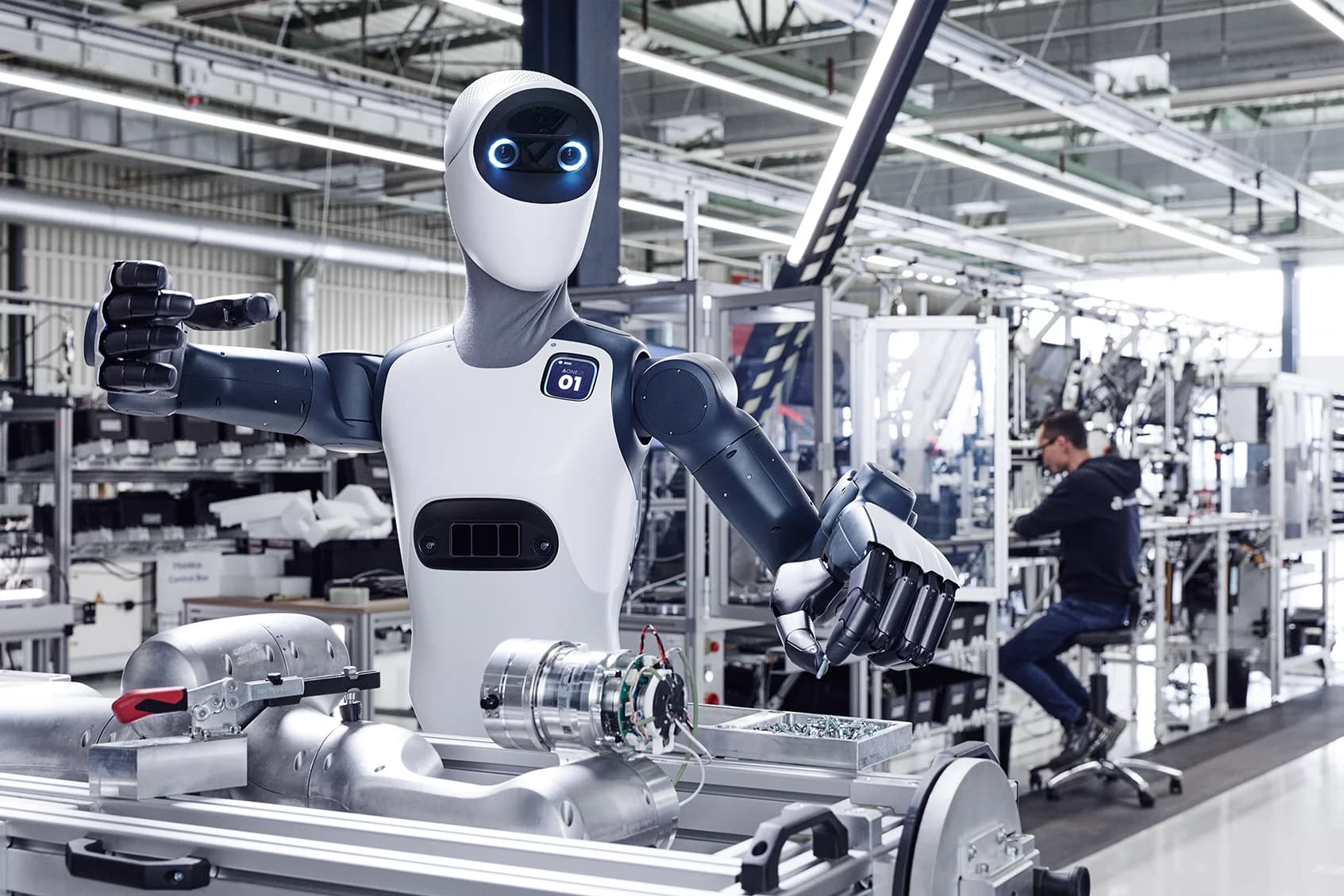 A waist-up view of the Agile ONE robot on a factory floor, gesturing with its black dexterous hands while a human worker operates machinery in the background.