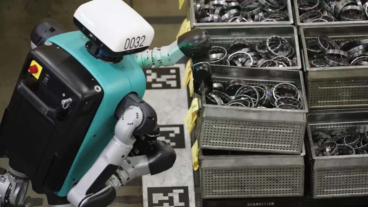 A top-down, side-view of an Agility Digit humanoid robot with a teal torso and a white head numbered 0032.  The robot reaches into one of several stacked metal bins, which are labeled SCHAEFFLER and filled with small, metallic bearing rings.  Black and white fiducial markers for navigation are visible on the floor around the robot's base. 