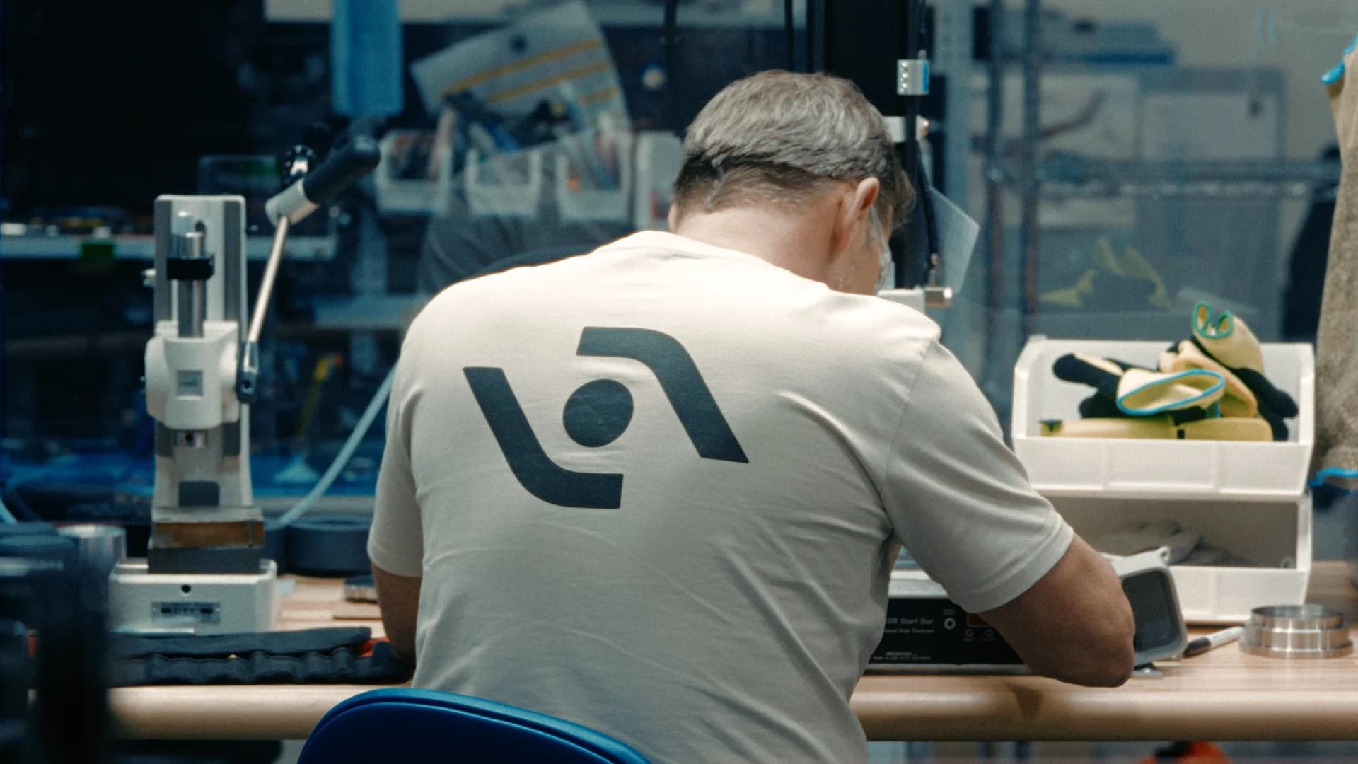 A man viewed from behind sits at a workbench in a manufacturing or workshop setting, wearing a tan t-shirt that displays the new Agility symbol in black on the back.