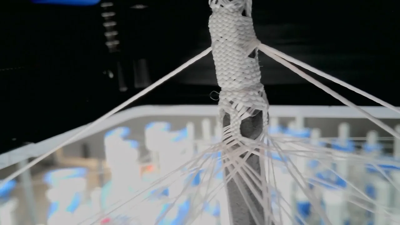 A macro shot of the Allonic braiding machinery in action, weaving white fibers directly around a dark skeletal core to form a robotic component.