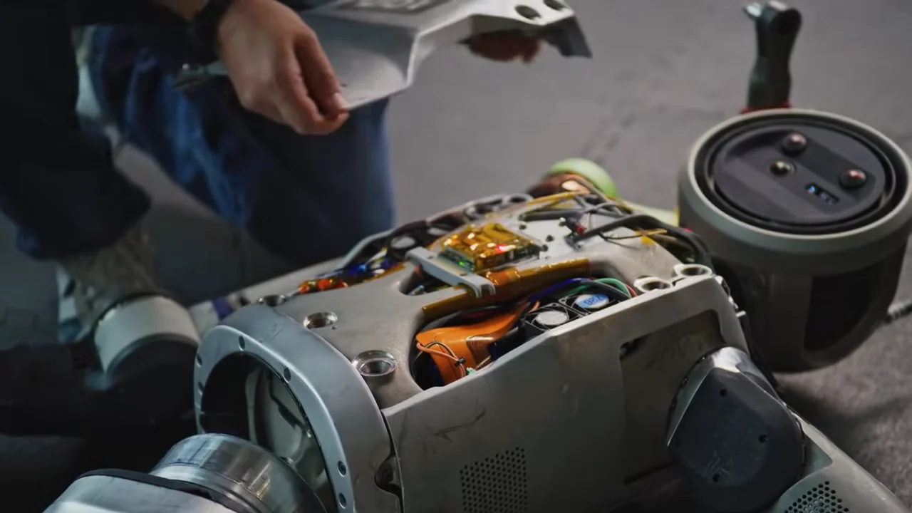 A Boston Dynamics Atlas robot lying supine on a laboratory floor with its chest chassis removed, exposing a complex array of internal cabling and electronics. A technician, visible only by a hand and knee, is kneeling beside the robot to perform maintenance on the hardware.