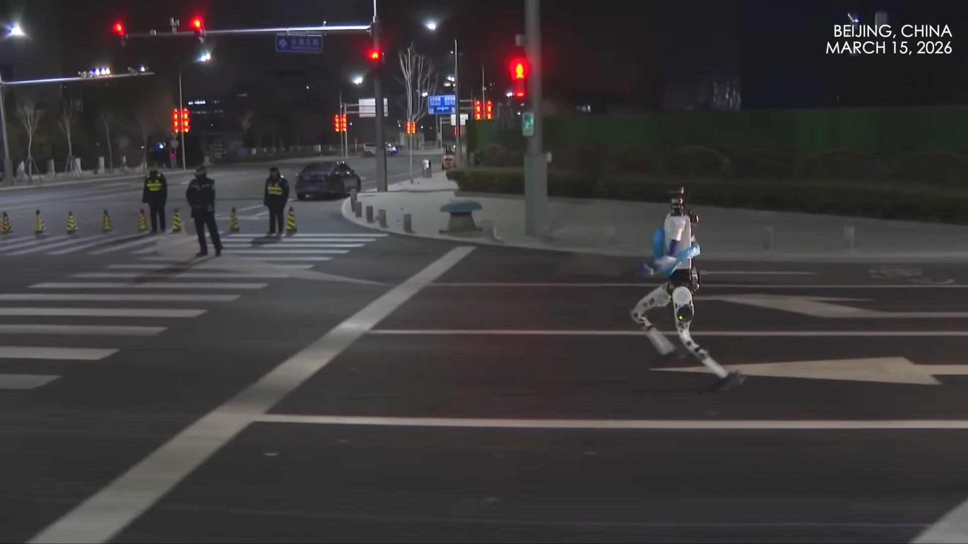 A white and blue Tiangong humanoid robot sprinting across a multi-lane city street in Beijing at night. The robot is captured in a clear flight phase with both feet off the ground, set against a backdrop of traffic lights and personnel in high-visibility vests.