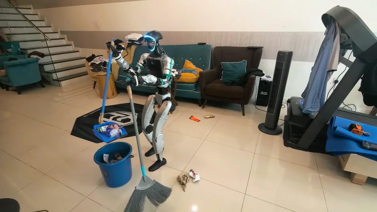 A silver and black Unitree G1 humanoid robot stands on a tiled living room floor, holding a long-handled grey broom to sweep up crumpled trash. A blue plastic bucket sits on its left containing paper debris, and a blue dustpan holding discarded packaging rests nearby. The background reveals an unscripted domestic setting with a teal sofa, a brown armchair, scattered clothing, and a treadmill.