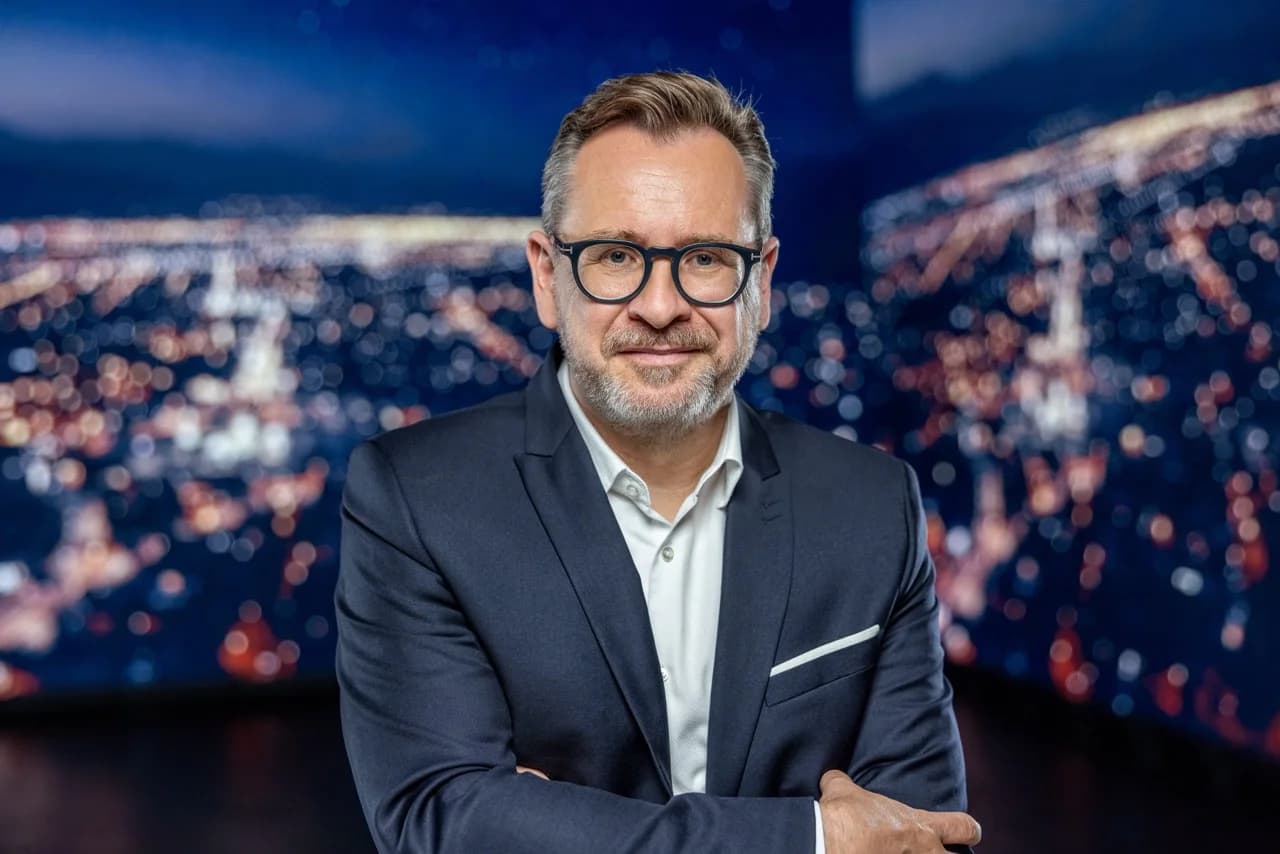 A professional portrait of Burkhard Boeckem, CTO of Hexagon, wearing a dark suit and glasses against a blurred city skyline at night.