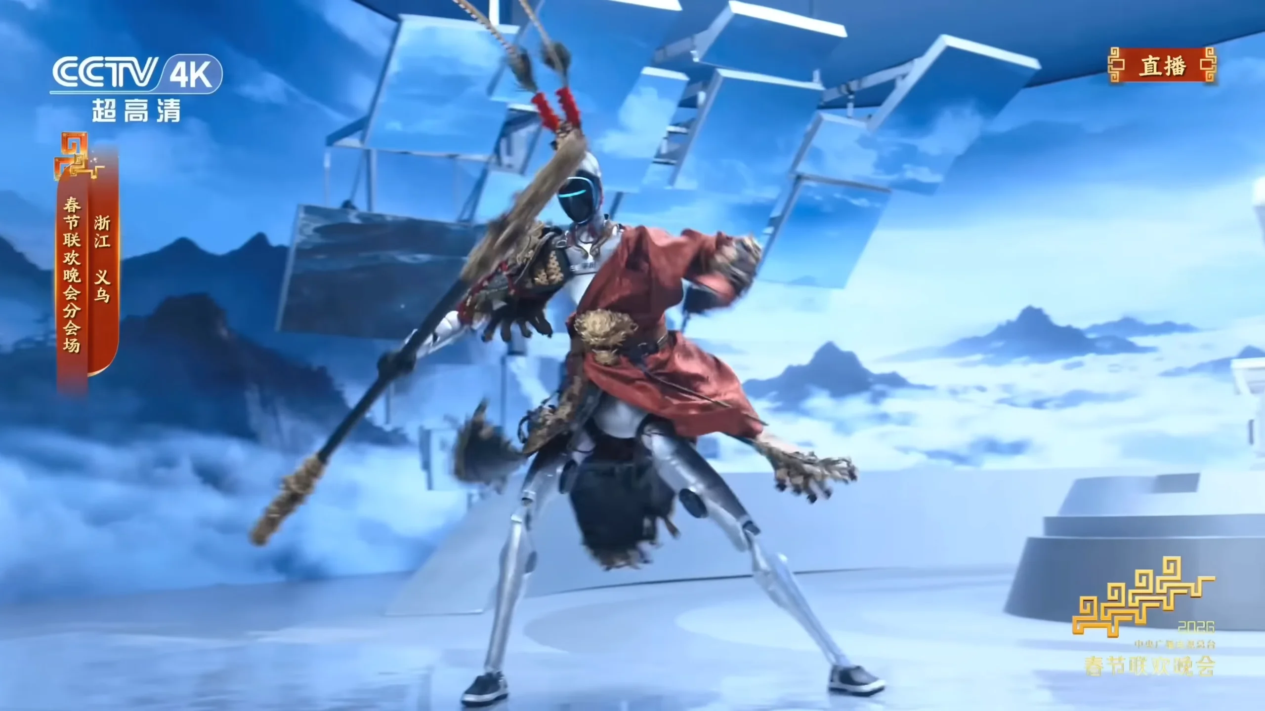 A large Unitree H2 humanoid robot dressed in a traditional red-and-gold warrior costume, performing a martial arts pose with a long staff. The background features a futuristic blue stage with mountain silhouettes and a CCTV 4K logo.