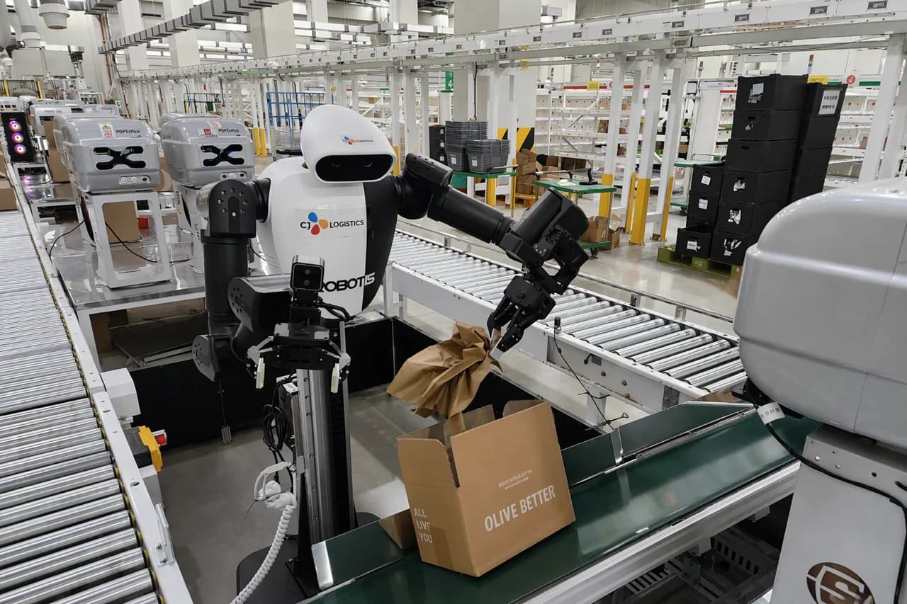 A dual-arm ROBOTIS AI Worker robot with a white torso and black arms standing at an industrial conveyor belt, holding crumpled kraft packaging paper with its mechanical gripper and placing it into an open cardboard box labeled 'OLIVE BETTER'.