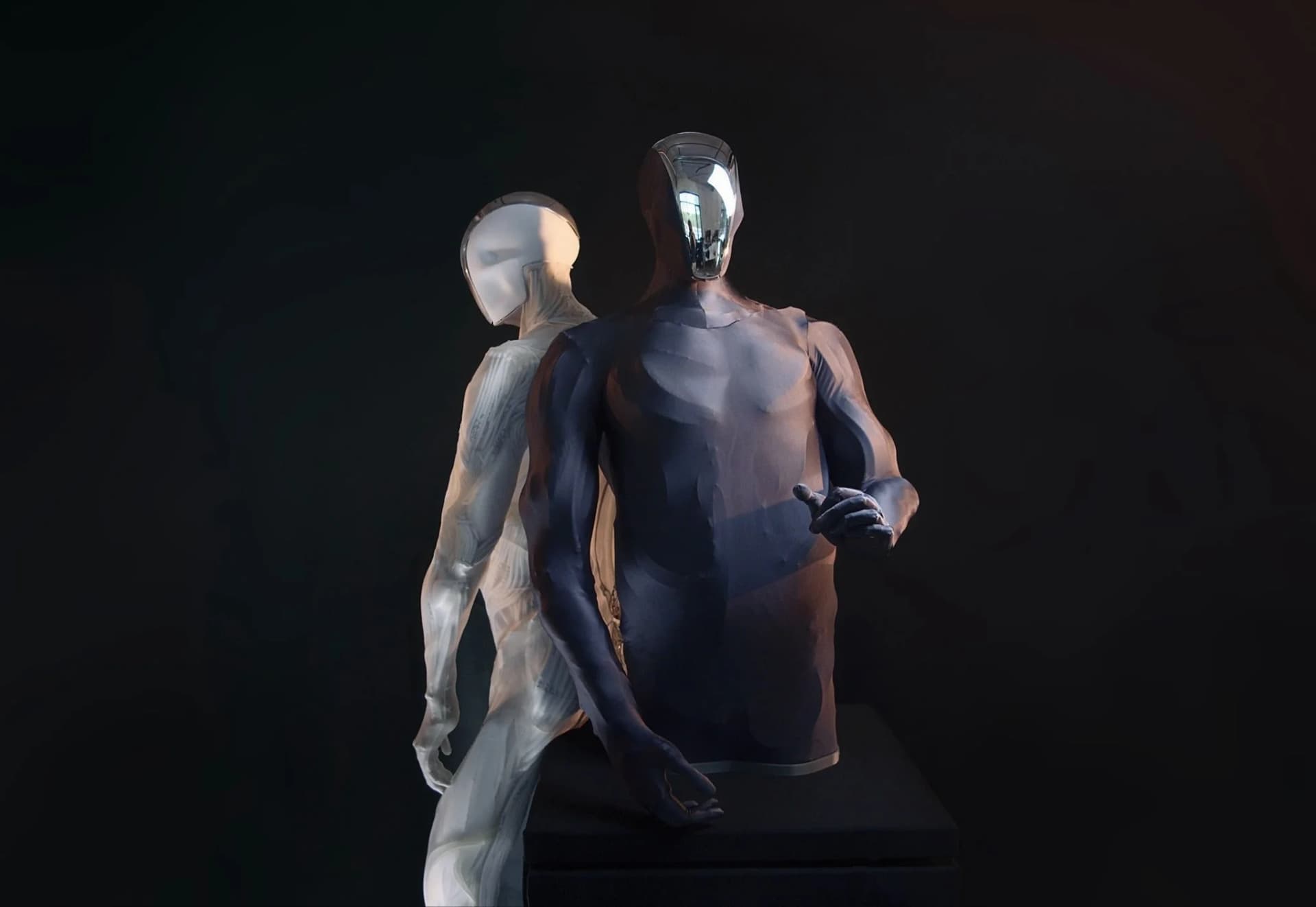 Two Clone robotic torsos shown back-to-back; one features a dark skin with a mirrored visor, while the other is translucent, revealing an intricate network of white artificial muscles.