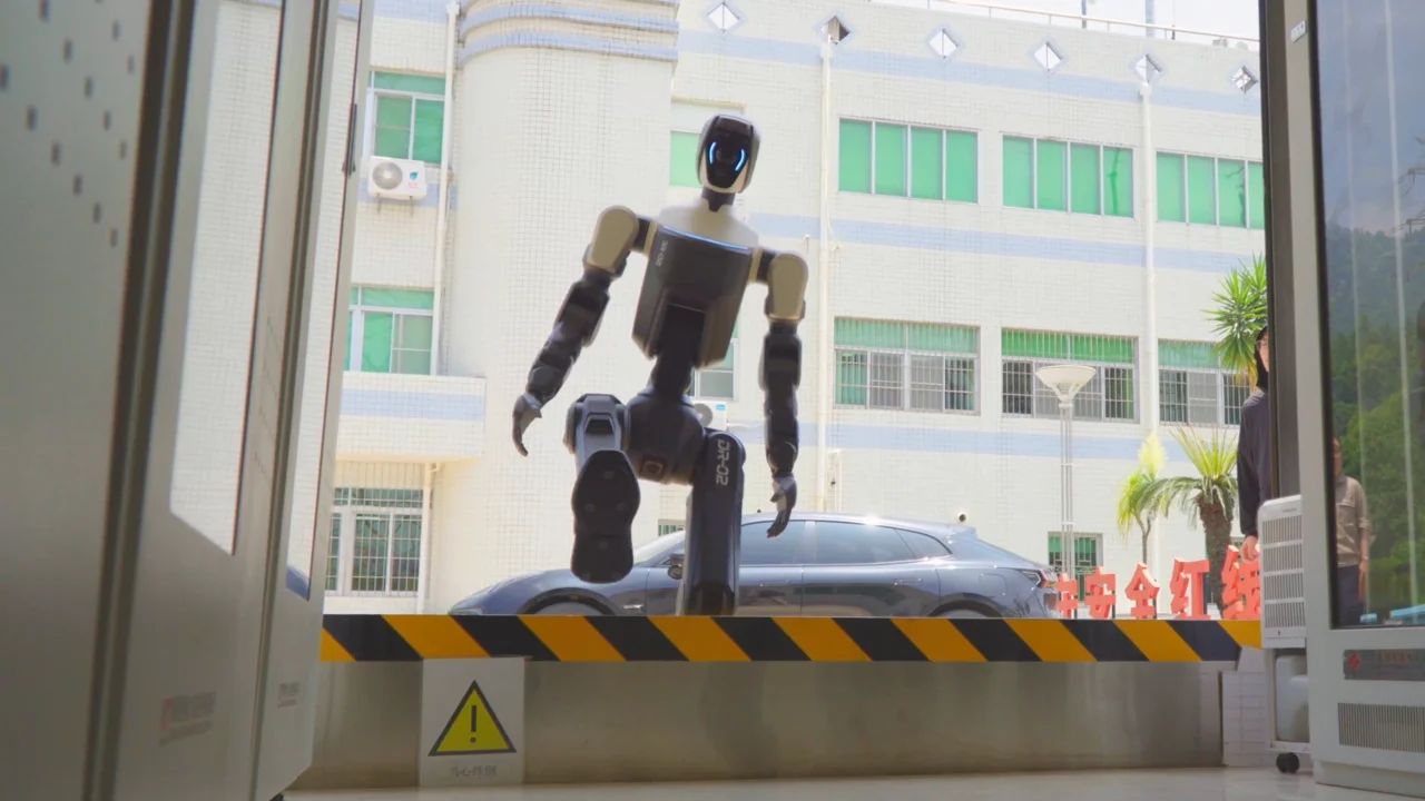 The Deep Robotics DR02 humanoid robot balancing on one leg as it steps over a raised yellow and black striped hazard barrier to enter an industrial facility. A building and parked car are visible in the background.
