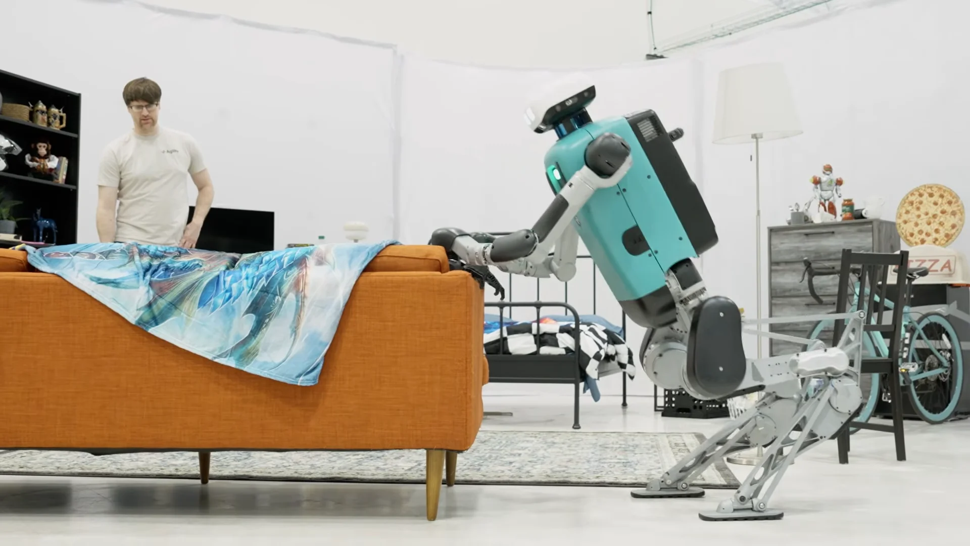 Agility's bipedal humanoid robot Digit leans forward and pushes a large orange sofa across a staged mock apartment inside a white-walled testing facility. In the background, a man in an Agility t-shirt looks on near a bed, a dresser, and a bicycle.