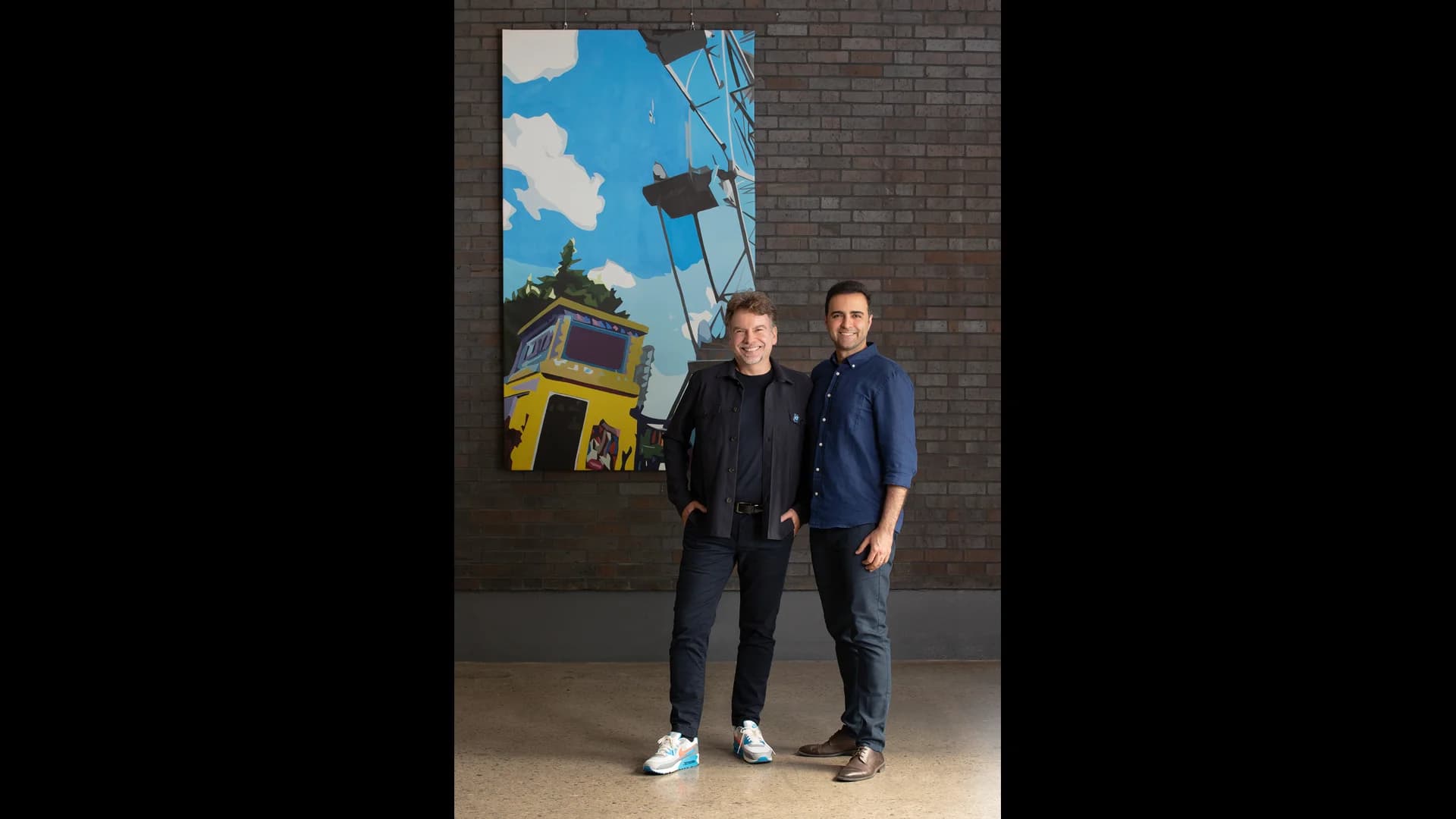 Dynamic Creatures co-founders Marc Theermann and Farbod Farshidian standing together in front of a dark brick wall with a colorful painting depicting a theme park Ferris wheel.
