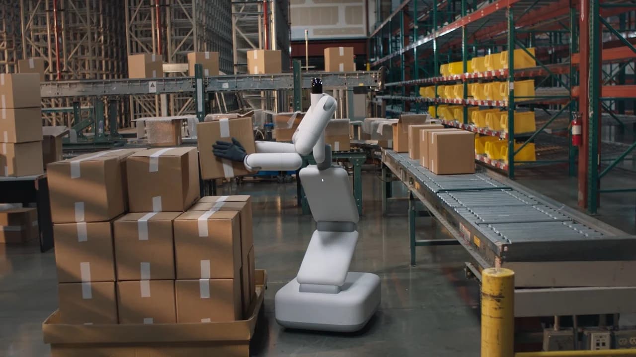 The Genesis AI Eno robot, featuring a wheeled base and an articulated white torso, uses its dual arms to lift a cardboard box off a pallet in a warehouse setting.