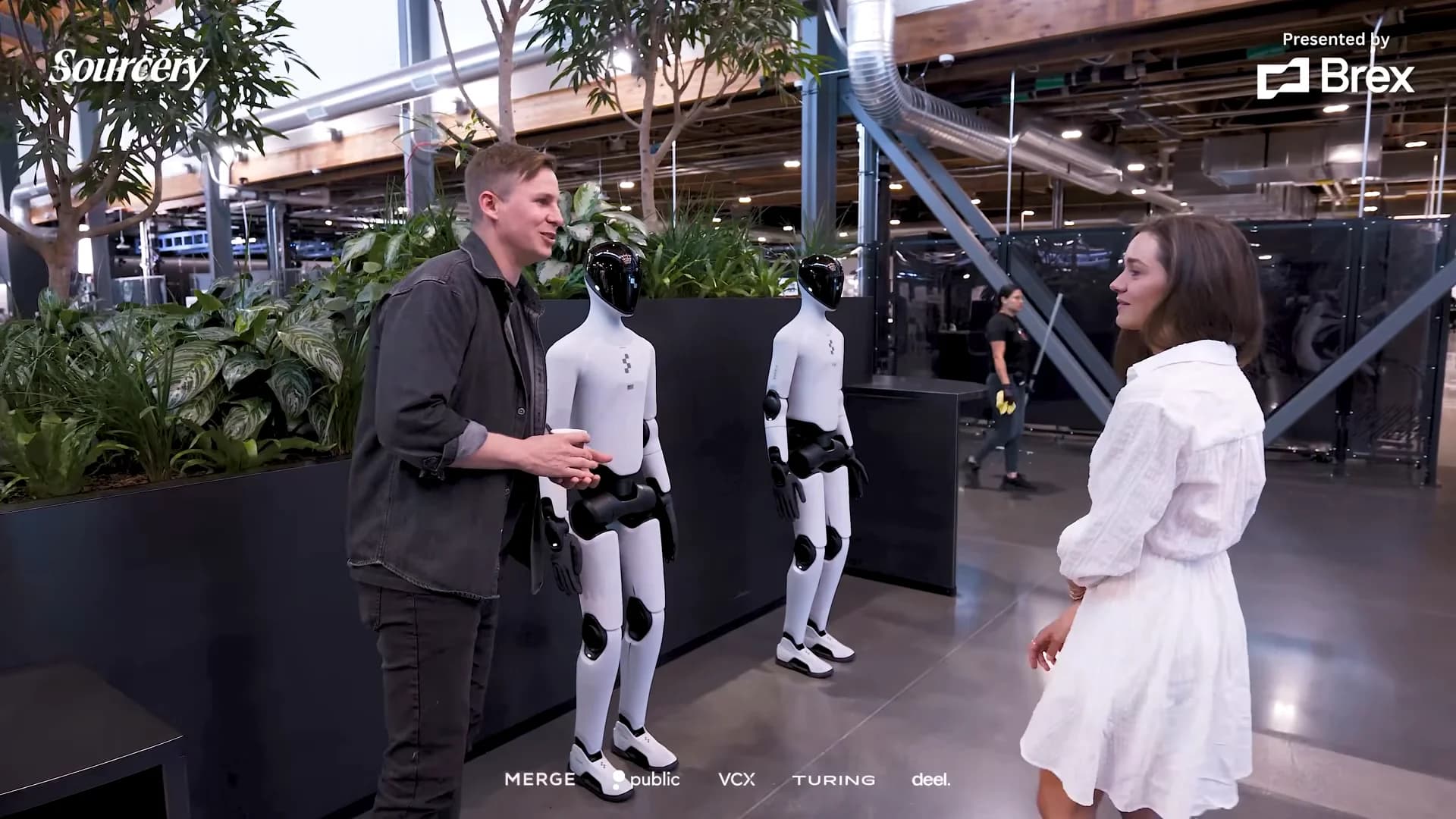 Inside Figure's headquarters, CEO Brett Adcock gestures toward two sleek white and black humanoid robots while speaking with interviewer Molly O'Shea. In the background, a human staff member is visible performing maintenance tasks.