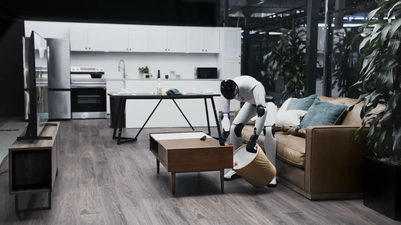 The Figure 03 robot leans over a wooden coffee table, holding a tan woven bin with its left hand while using its right hand to scoop colorful building blocks into the container.