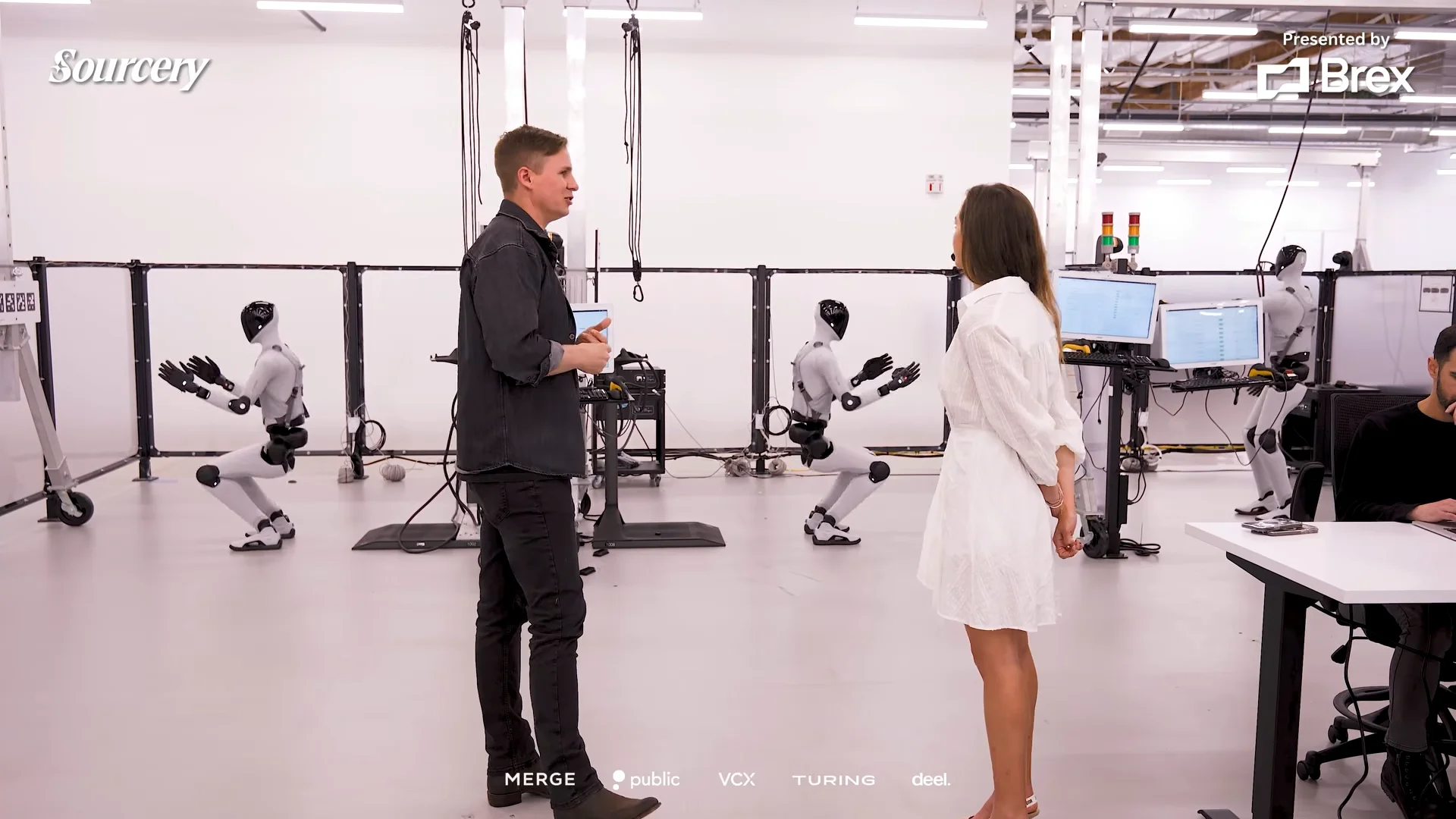 Inside Figure's system integration lab, Brett Adcock and Molly O'Shea stand amidst several humanoid robots performing repetitive deep squats within specialized testing bays.
