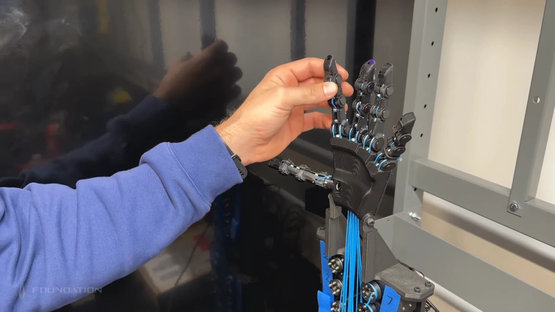 A close-up view of a robotic hand mockup featuring a complex system of bright blue strings acting as tendons. A person's hand is visible, manipulating the index finger of the prototype. The mechanical structure of the hand and the routing of the blue tendons into the forearm are clearly visible.