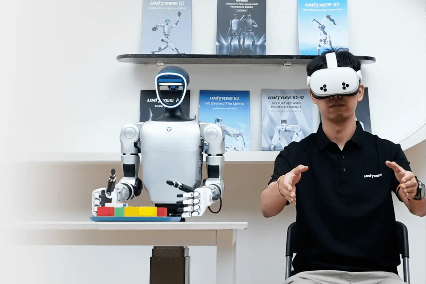 A researcher wearing a VR headset sits next to a G1-D robot, using hand controllers to teleoperate the robot's arms in real-time.