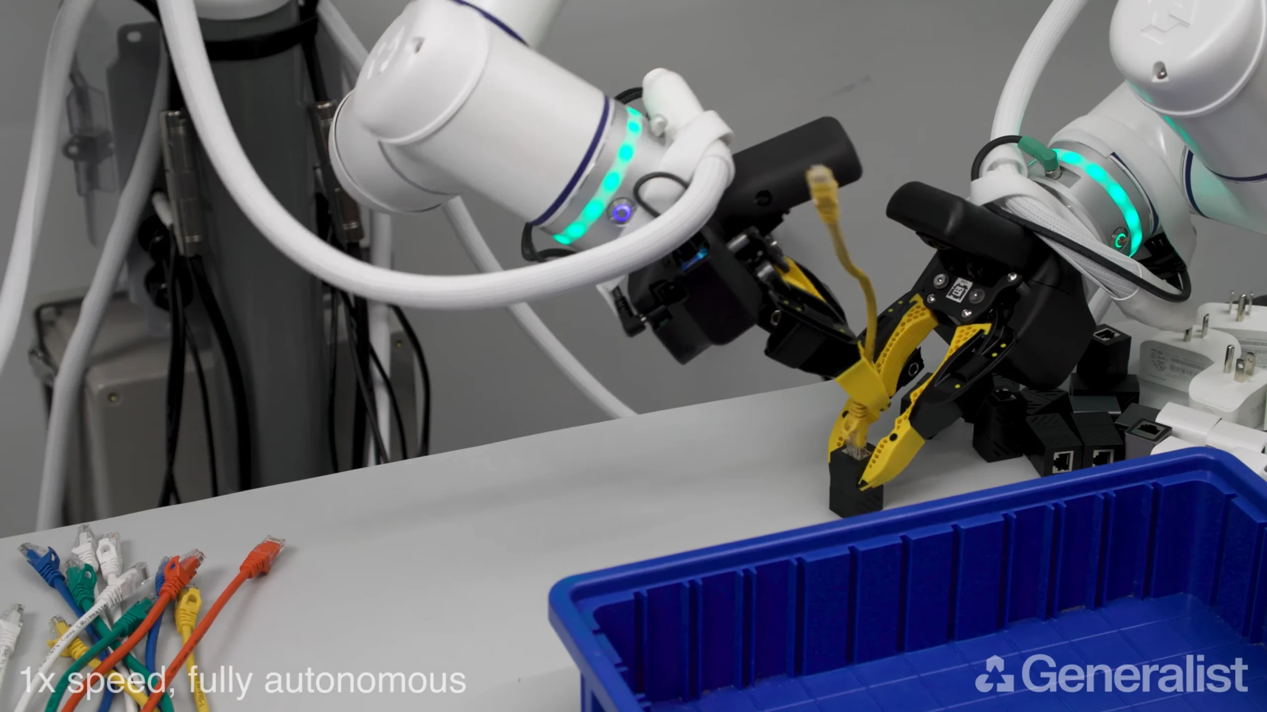 A high-angle screenshot of a Generalist AI robot with two white articulated arms. The arm on the right holds a black handheld ethernet socket, while the arm on the left, using a yellow-tipped gripper, plugs a yellow ethernet cable into the socket. A blue bin and multiple colored ethernet cables sit on the white workspace. Labels at the bottom indicate the operation is fully autonomous at 1x speed.