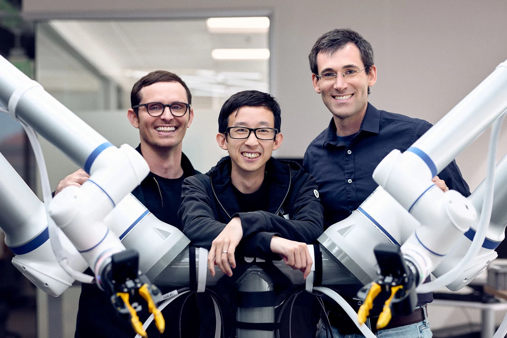 Three smiling men from the Generalist AI leadership team posing behind a dual-arm robotic system. The robot features two white articulated arms with blue bands and yellow grippers extending into the foreground. The man in the center, wearing glasses and a dark hoodie, leans comfortably on the robot's central support pillar.