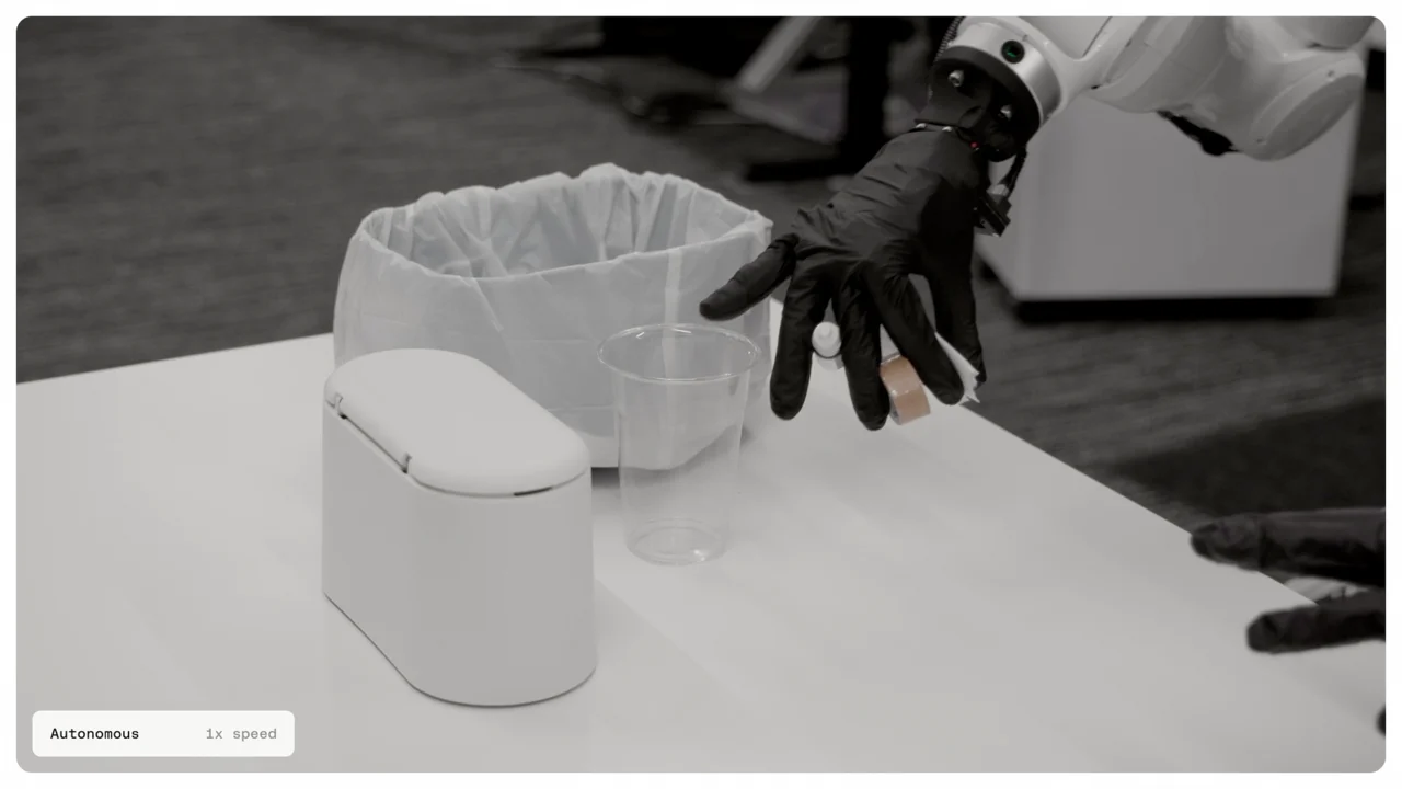 A dexterous robotic hand with a black glove simultaneously holding four small objects of varying sizes, including a roll of tape and a small bottle, over a white table.