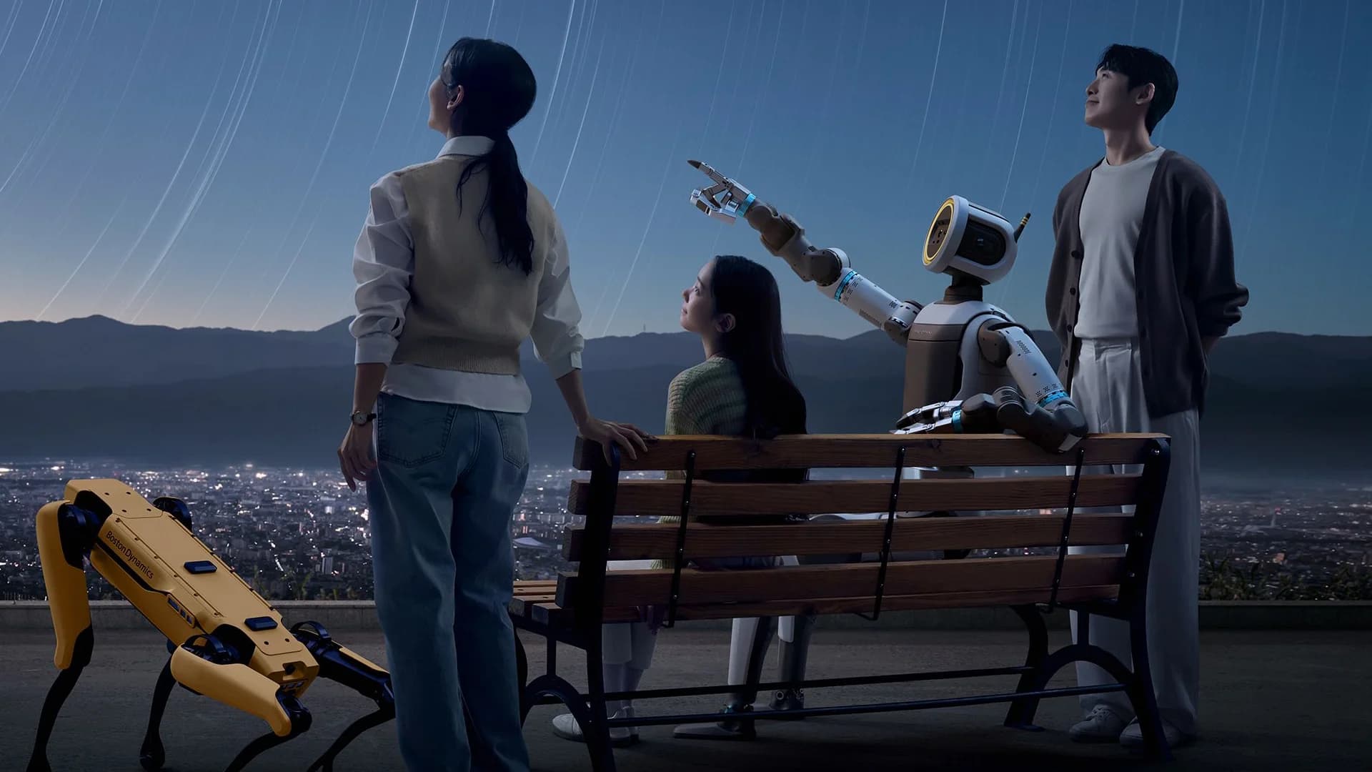 A cinematic shot of a yellow Spot robot and an Atlas humanoid robot on a park bench next to three people, all looking up at a night sky filled with star trails.