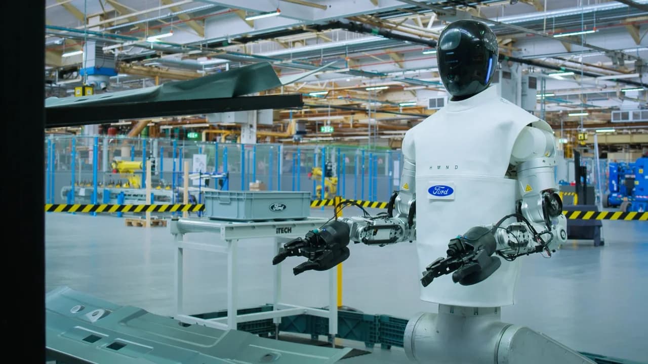 Branded with the Ford logo, a HMND 01 Alpha robot  wearing a protective white garment  operates in the Ford Innovation Centre in Cologne, positioned near metal car body components.