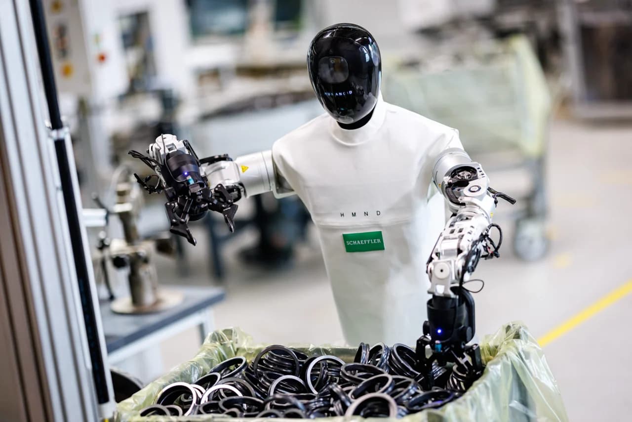 A HMND 01 Alpha robot uses parallel grippers to pick metallic bearing rings from an industrial bin. The robot is fitted with a white fabric uniform featuring a green Schaeffler patch.