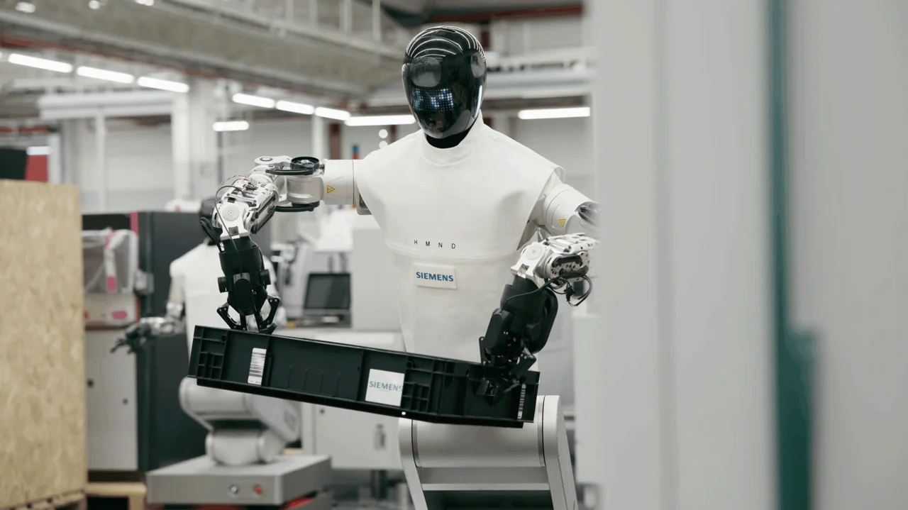 A wheeled HMND 01 Alpha robot , wearing a white fabric 'uniform'  with a Siemens logo, carries a black industrial tote through the Siemens Electronics Factory in Erlangen.