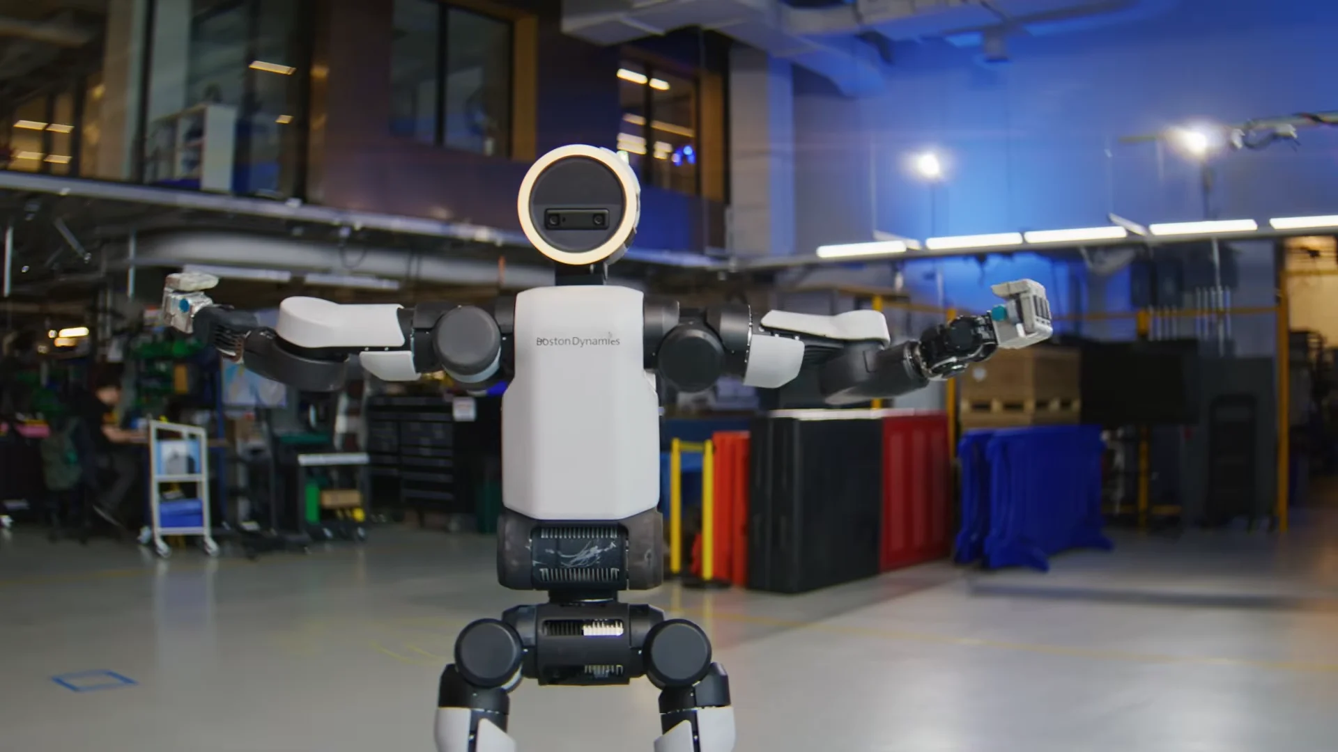 A front-facing view of the all-electric Boston Dynamics Atlas robot standing centered in a workshop with its arms extended fully to its sides and its circular head illuminated by a white ring light.