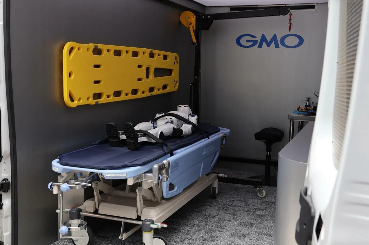 Interior of the GMO Humanoid Ambulance showing a small humanoid robot strapped onto a blue medical gurney next to repair tools and an engine hoist