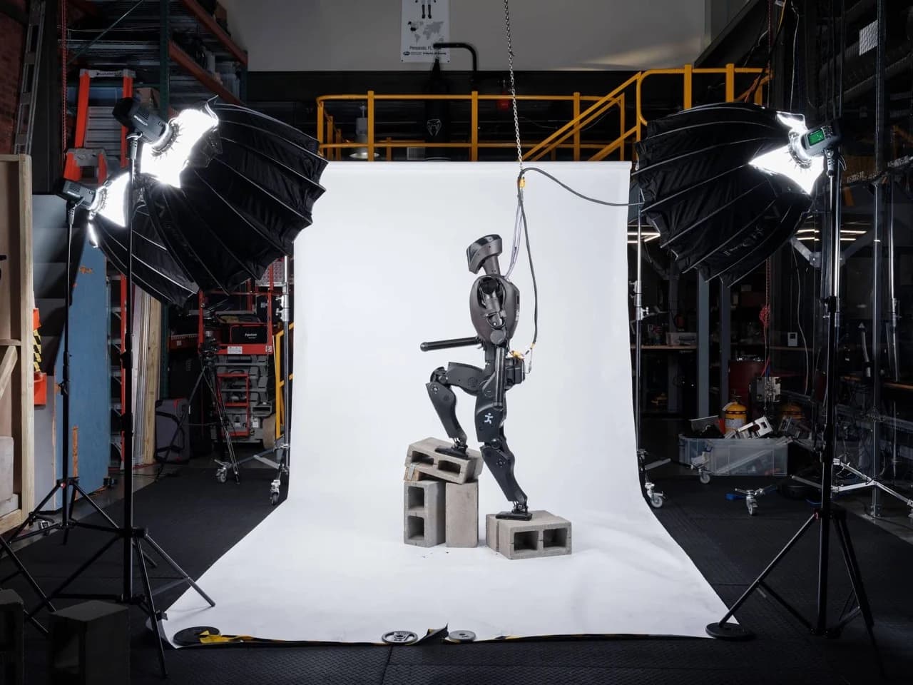 A profile shot of the IHMC research robot, Alexander, standing on concrete blocks in a studio or laboratory setting. The robot is charcoal black with a humanoid form and is tethered from above. Professional studio lighting and photography equipment are visible in the background.