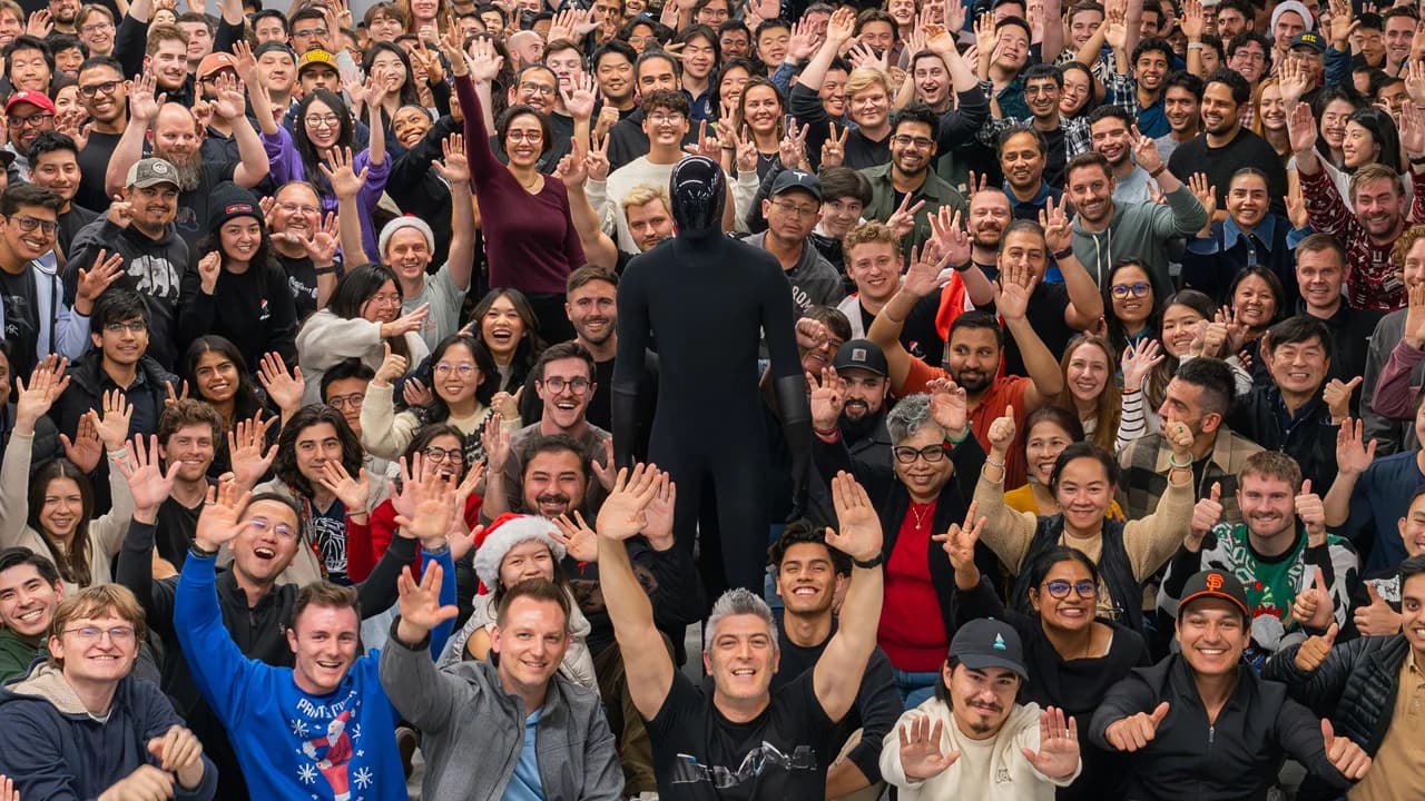A large group of smiling Tesla Optimus team members waving and posing for a group photo. In the center stands a sleek, faceless, all-black humanoid robot. Several people in the crowd are wearing festive holiday items, including a Santa hat and Christmas sweaters.