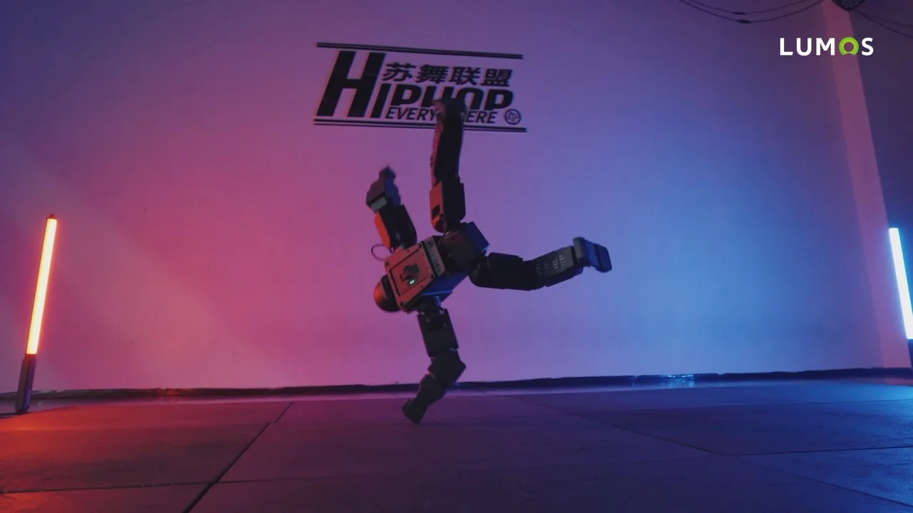 A compact, black humanoid robot executing a dynamic breakdancing freeze on one arm in a studio with dramatic red and blue stage lighting.