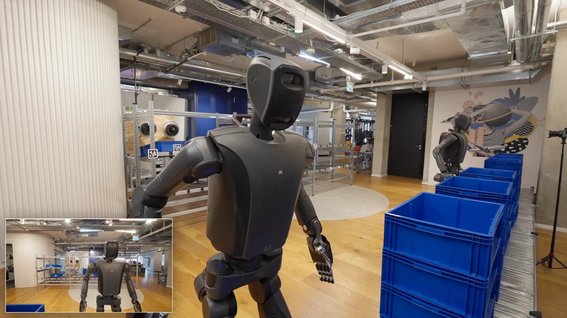 A screenshot from a Mentee Robotics video showing two dark grey humanoid robots in a well-lit, simulated warehouse environment with wooden floors. In the foreground, a V3 robot walks past stacks of blue plastic crates, while a second robot stands near metal shelving in the background. A small picture-in-picture camera view is visible in the bottom left corner.