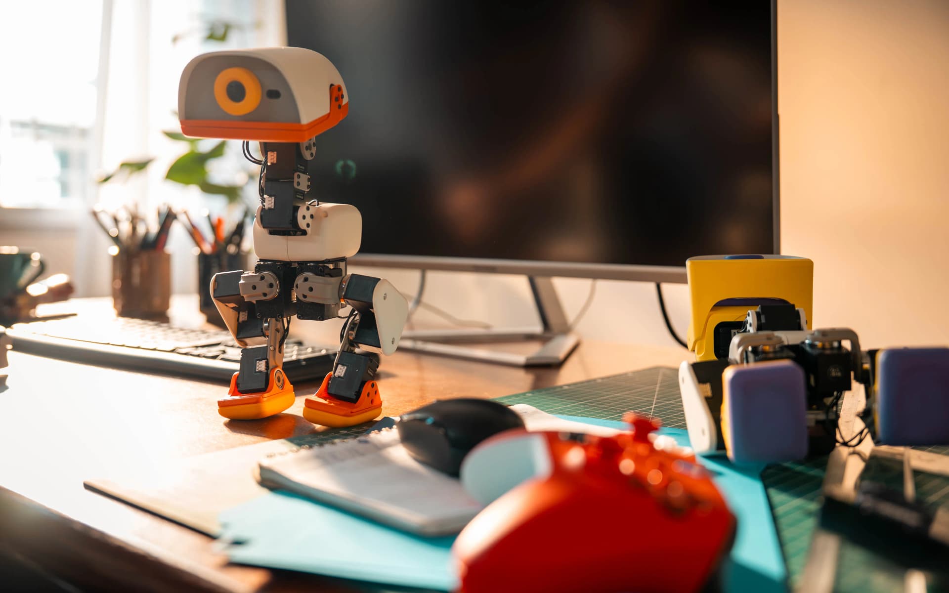 A Cream colourway Microduck bipedal robot standing on a wooden desk in morning light in front of a computer monitor, beside a game controller and parts from other units.