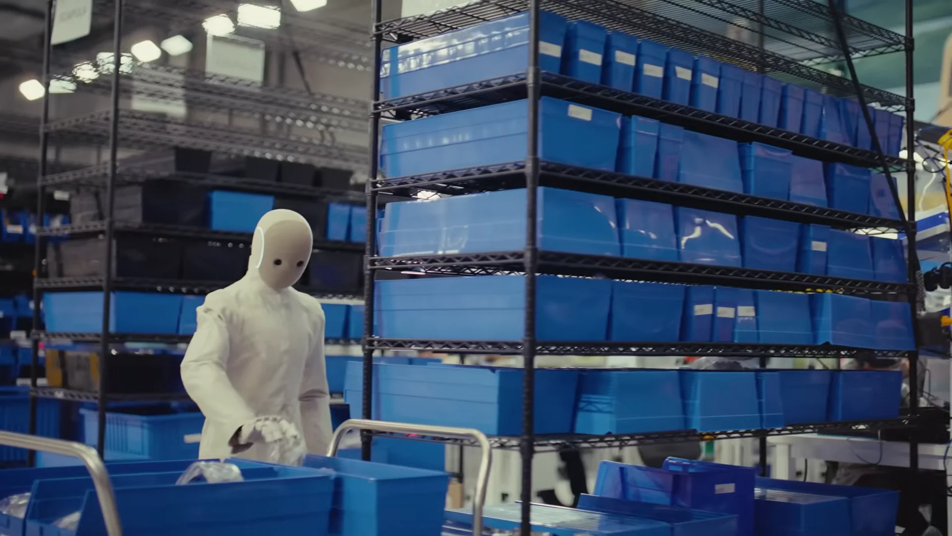 A white, textile-clad NEO humanoid robot standing in a factory aisle, reaching into a blue plastic storage bin on a rolling cart next to tall industrial shelving units.