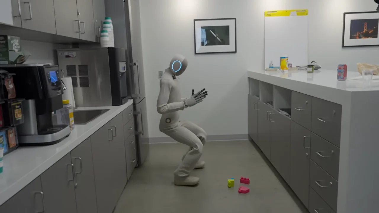 1X NEO humanoid robot attempting to squat down to pick up a tong from the floor in the Hard Fork podcast
