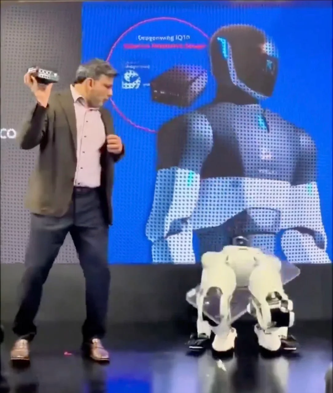 A screenshot showing Qualcomm executive Nakul Duggal speaking on stage and holding a compute module. In the foreground, a NEURA Robotics 4NE-1 humanoid has collapsed forward onto its knees.