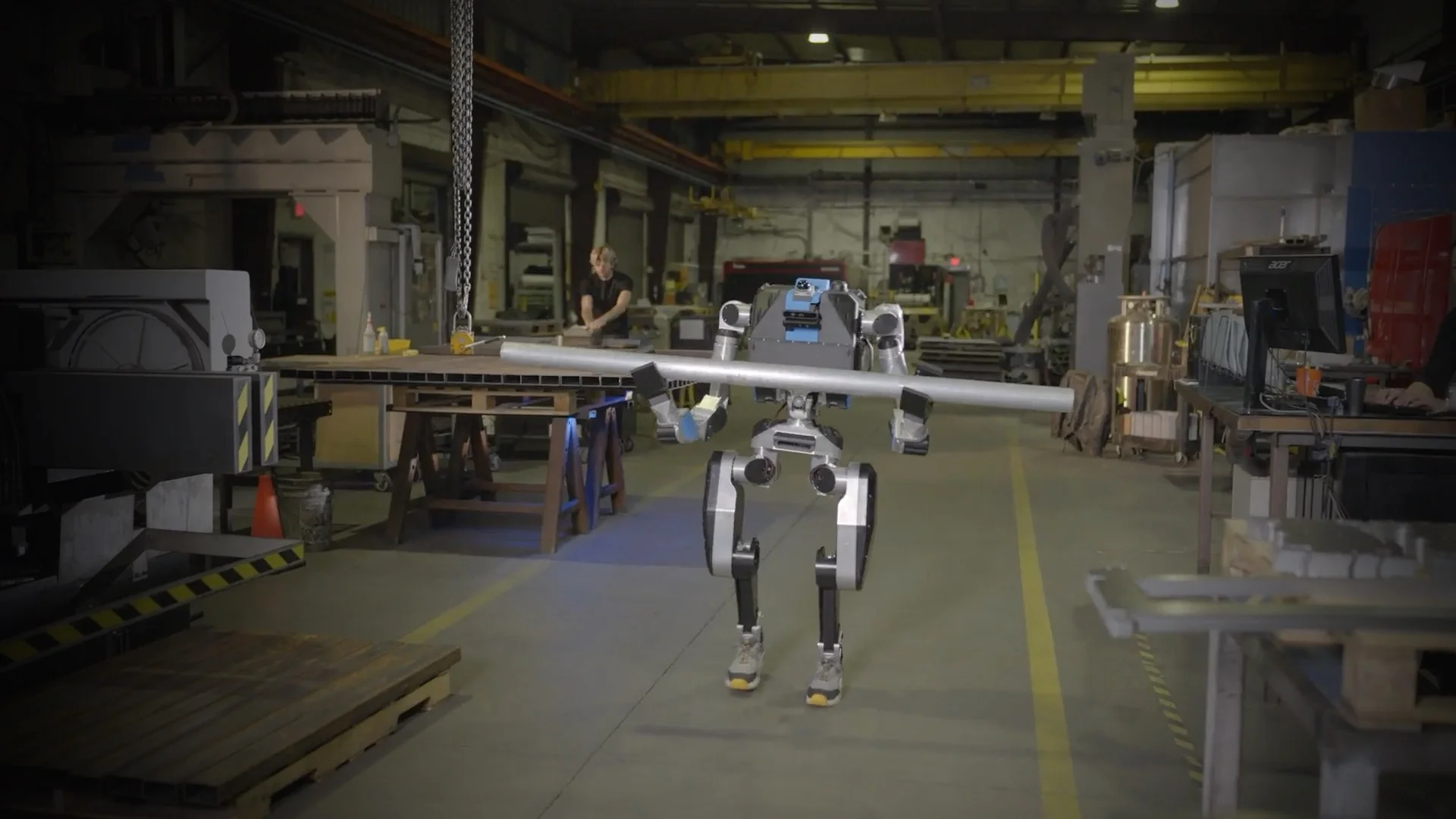 A bipedal Noble Machines robot carrying a long metal pipe horizontally through an industrial facility.