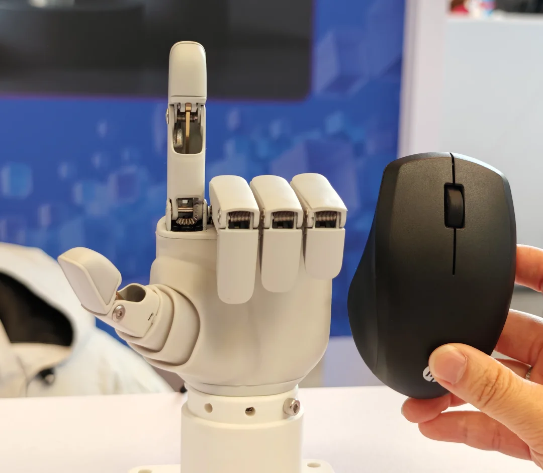 A close-up comparison showing a compact white robotic hand with its index finger extended upward, positioned next to a standard black computer mouse held by a human hand. The image highlights that the robotic hand is roughly the exact same size as the mouse.