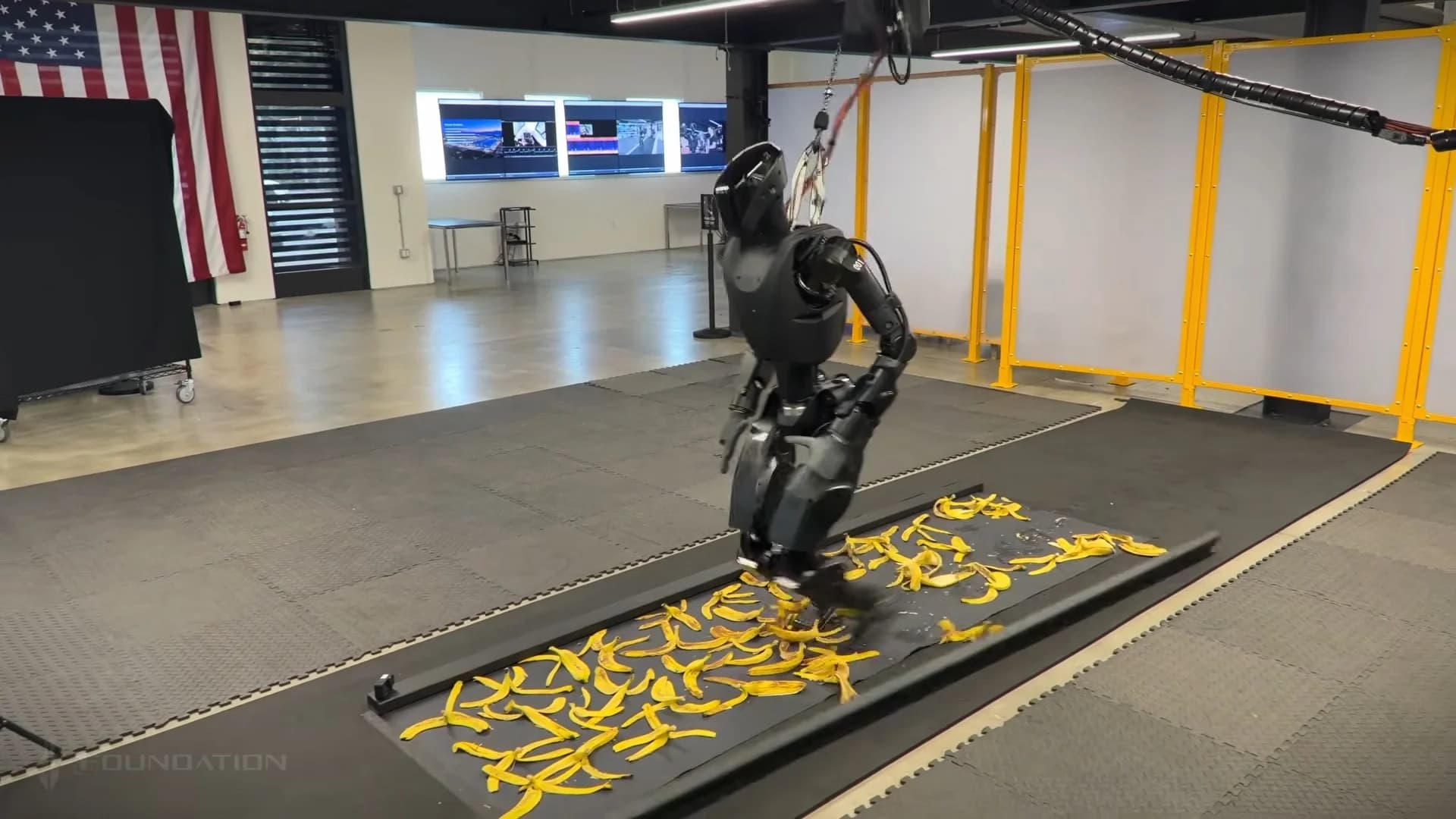 A charcoal-black Foundation Phantom humanoid robot attempts to walk across a floor covered in yellow banana peels while tethered to a safety gantry. The robot is shown mid-stride in a laboratory setting with its internal actuators and structural rods visible.